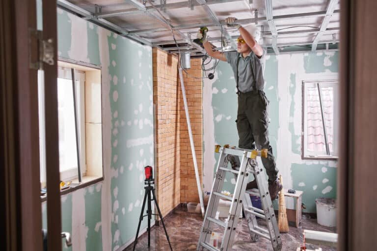 Ouvrier en tenue de sécurité installant des câbles électriques sur une armature de plafond suspendu dans une pièce en rénovation avec des murs partiellement enduits et une fenêtre ouverte, reflétant une phase active de travaux d'électricité.