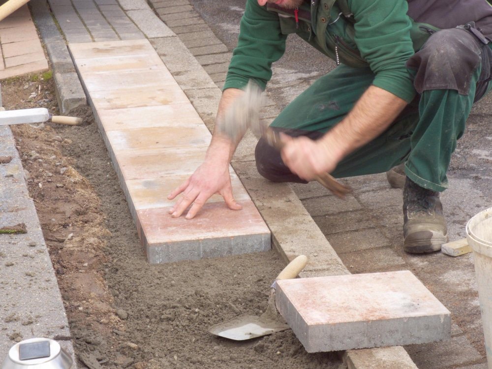 Ouvrier en tenue de travail posant des dalles de trottoir avec précision, en mouvement dynamique, sur un chantier de paysagisme extérieur.