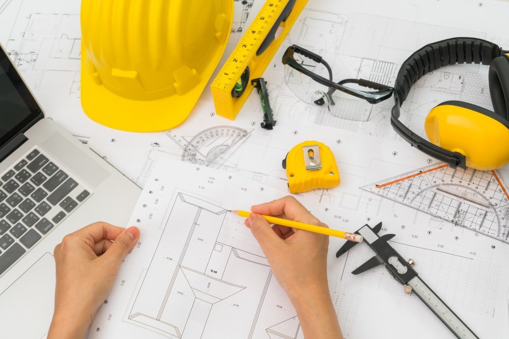 Espace de travail d'architecture avec des plans étalés sur une table, un casque jaune, des lunettes de protection, et des écouteurs, avec une personne tenant un crayon et travaillant sur les détails du projet.