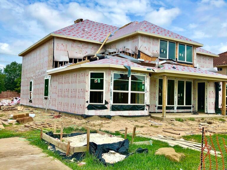 Maison résidentielle en phase de gros œuvre avec isolation extérieure et toit partiellement complété, entourée de débris de construction sur un terrain en herbe.